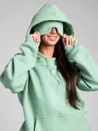 Person wearing a light green hoodie with the hood covering their eyes on a gray background