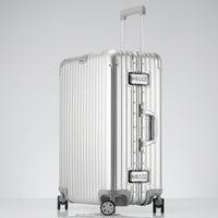 A three-quarter front-angle studio view of The Masterpiece — The Legacy aluminum-magnesium alloy suitcase in a brushed Sterling Silver finish. The telescopic handle is partially extended, and the grooved, zipperless metallic shell is visible along with its dual integrated TSA-approved locks and side handle. The luggage stands on four multi-directional spinner wheels against a neutral studio background. No external branding is present on the casing.
