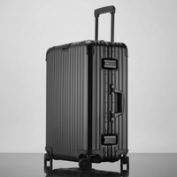 A black ribbed aluminum spinner suitcase with an extended telescopic handle, standing upright on a reflective dark surface against a light gray studio background.