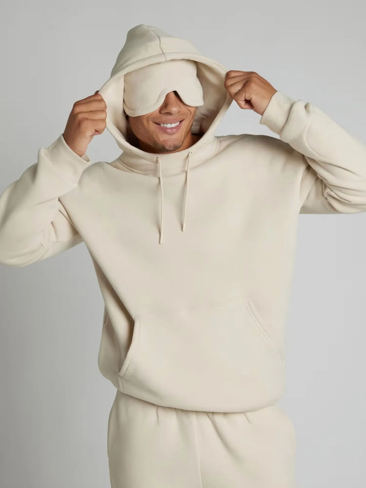 Person wearing a beige hoodie and matching pants with a sleep mask on a plain background