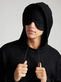 Person wearing a black hoodie and eye mask against a gray background
