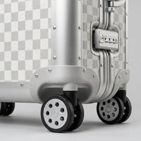 Close-up detail of Aeria Prestige The Monarch aluminum travel case showcasing smooth spinner wheels, reinforced corner riveting, and an integrated TSA-approved combination lock in Alabaster White Damier finish.