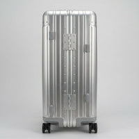 A full-length front view of The Masterpiece — The Legacy aluminum-magnesium alloy travel case in a Sterling Silver finish, displayed against a neutral studio background. The suitcase features a zipperless design with heavy-duty reinforced metallic hinges and rivets. The shell showcases a metallic, grooved silhouette, and four silent multi-directional spinner wheels are visible at the base.